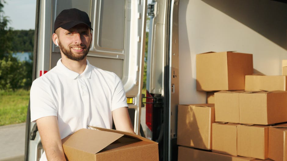 A man with a beard and wearing a black cap and white polo shirt is standing outside near an open moving van, holding a cardboard box. The van’s interior is visible, with additional boxes neatly stacke