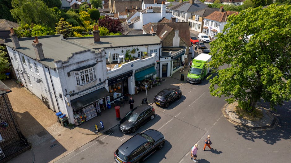 An aerial view of a busy street in Bromley Common showing a small, white, two-storey shop with large windows and a green awning; adjacent to the shop, there are several parked vehicles including a green van, a black car, and a grey van, with people walking on the pavement and near the vehicles. Behind the shop, residential houses with tiled roofs and trees with green foliage are visible, indicating a suburban area suitable for home relocation and furniture transport. The street is partly shaded by trees, and the scene captures the typical moving logistics involved in house removals, such as loading and parking arrangements for a local man with van service, with no visible moving activity happening at this moment.