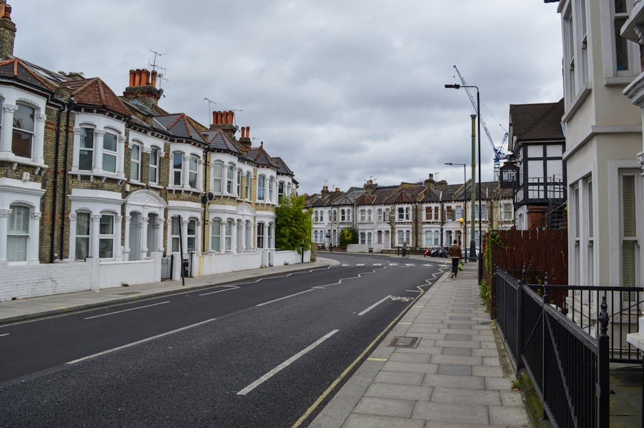 A quiet residential street in Bromley Common featuring a row of Victorian-style terraced houses with white facades, bay windows, and red brick chimneys, lined along a narrow, one-lane road paved with asphalt. The pavement on the right side of the image is made of concrete slabs, with black metal railings bordering the front gardens of the houses. Overhead, there are grey, cloudy skies, contributing to an overcast atmosphere. Streetlights and a tall construction crane are visible in the background, indicating ongoing development or building work. The street appears to be part of a typical home relocation or furniture transport route, with no moving activity currently visible, but the scene reflects the environment where a professional removals service, such as Man with Van Bromley Common, could operate during house removals or moving procedures.