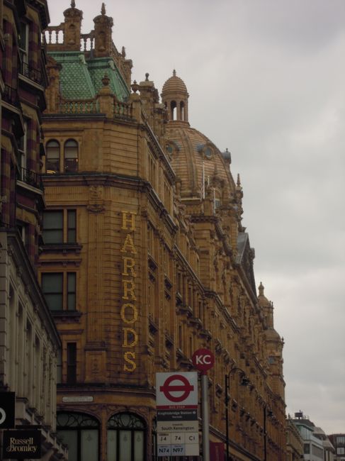 The image shows the ornate exterior of a historic building with intricate architectural details and a prominent sign reading 'HARRODS' on the facade, located next to a busy urban street in Bromley Com