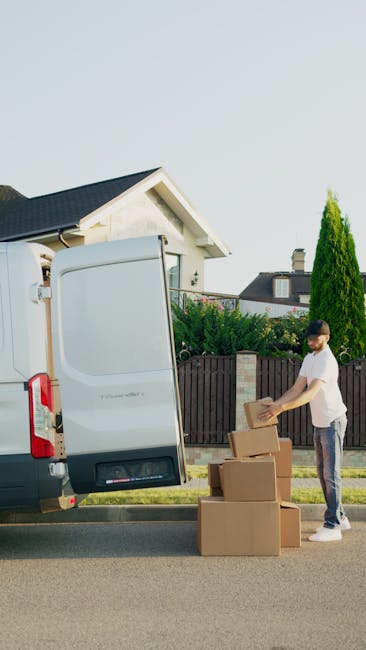A man stands outside a residential property onto a paved street, preparing to load or unload cardboard boxes from the rear of a white van parked on the roadside. The van has its rear doors open, revealing an interior used for packing and moving furniture or household items. The man is placing a cardboard box onto a small pile of similar boxes on the pavement, with additional boxes stacked nearby. In the background, there is a single-story house with a pitched roof, complemented by a garden featuring a tall, slender evergreen tree and other greenery. The scene is set in daylight with clear skies, consistent with an efficient home relocation or furniture transport process, and is representative of professional removals services offered by companies like Man with Van Bromley Common.