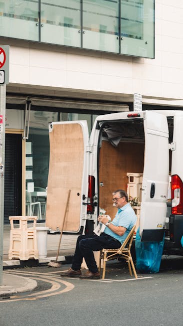 A man wearing glasses and a light blue shirt is seated on a wooden chair beside a white delivery van parked on a city street, demonstrating a break or planning during a home relocation or furniture transport process. The van's rear doors are open, revealing a partially loaded interior with large cardboard boxes, wooden panels, and packing materials, such as bubble wrap and plastic covers, arranged inside to protect furniture and household items during transit. The van is positioned close to a storefront with large glass windows and a modern building façade above, indicating an urban environment. Nearby, a wooden stool and a small plastic crate are placed on the pavement, possibly used for organizing items or transporting smaller belongings. The scene captures the loading or unloading process typical of professional removals, with a focus on careful handling and organized packing of household belongings, consistent with services offered by Man with Van Bromley Common in their house removals and moving logistics operations.